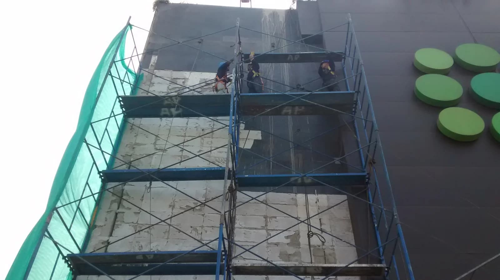 Cuadrilla con arnés sobre andamio metálico contra la fachada alta del Tottus San Luis junto al revestimiento de paneles oscuros y círculos verdes del logo TOTTUS