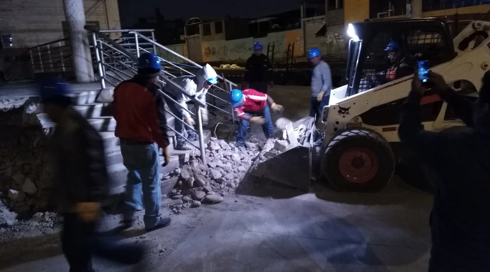 Cuadrilla con casco azul demoliendo rampa de concreto junto a escalera principal del ingreso de Tottus Quilca con Bobcat S650 cargando escombro de madrugada
