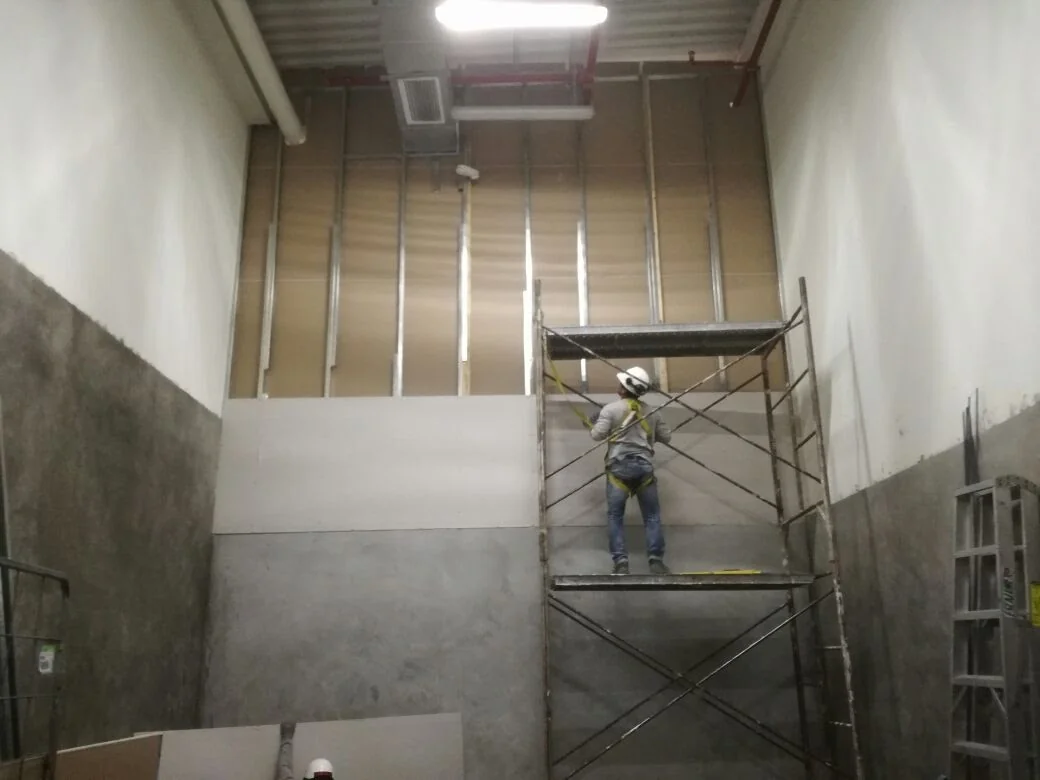 Operario sobre andamio con arnés instalando placas de drywall sobre estructura metálica galvanizada en falso muro de doble altura en almacén de Tottus Sullana